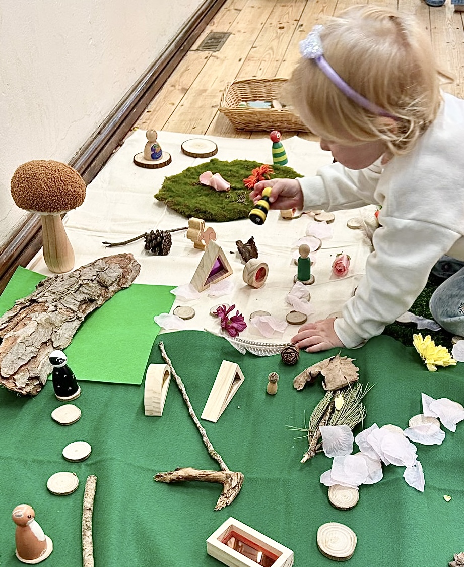 A child exploring peg people, bark, felt flowers and a bee puppet at a small-world invitation